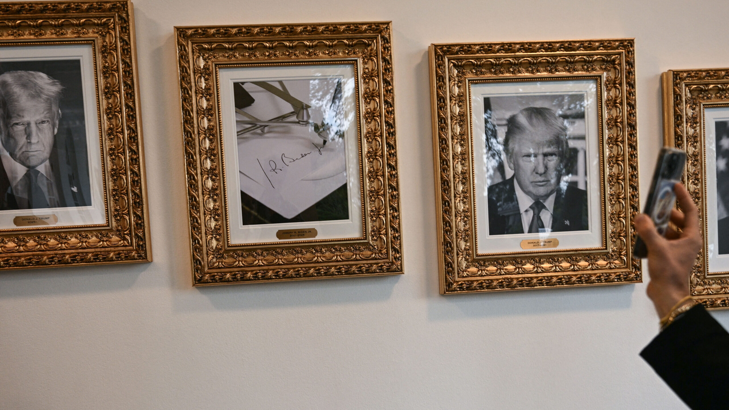 A hand holds a smartphone to photograph a row of framed portraits and a signed document hanging on a white wall.
