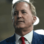 Ken Paxton, wearing a blue plaid blazer and a red tie, looks toward the side in this media portrait.