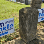 Two political campaign signs for Tom Perriello for Congress placed on a grassy lawn near a concrete post.