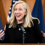 Abigail Spanberger with long blonde hair speaks and gestures at a podium, smiling. She wears a black outfit. U.S. and Virginia state flags are visible in the background.