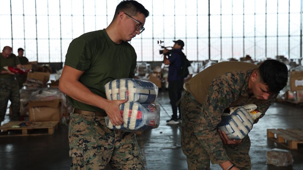 Two service members in camouflage uniforms carry packages of supplies in a large hangar.