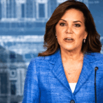 Michele Tafoya in a blue jacket, speaking at a podium flanked by two people.