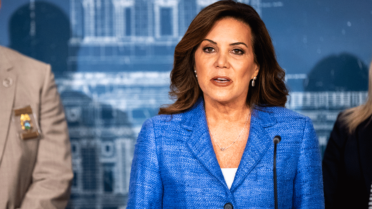 Home 1 Michele Tafoya in a blue jacket, speaking at a podium flanked by two people.