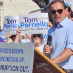 Tom Perriello speaking at a podium before a crowd holding Tom Perriello for Congress signs and a sign that reads PRICES DOWN. PAYCHECKS UP. CORRUPTION OUT.