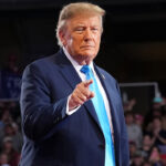 Donald Trump in a navy blue suit and bright blue tie pointing toward the camera at a freedom rally.
