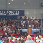 President Donald Trump speaks from a podium with the slogan Make America Great Again on a poster behind him. He speaks at a crowded outdoor rally in Georgia.