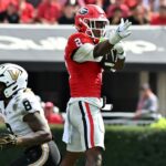 A Georgia football player in a red jersey catches a pass while a Vanderbilt player in a white jersey defends nearby.