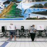 People vote at booths in a gymnasium with a large mural of Virginias map and landmarks on the wall. Some voters are standing, while others are walking, all engaged in the voting process.