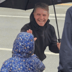 A man in a black jacket smiles while giving a high-five to a child in a blue floral jacket under an umbrella.