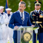 Pete Hegseth in a suit speaks at a podium with a Department of War seal, flanked by members of the military in uniform.