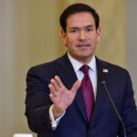 Marco Rubio speaks at a podium with American and Greek flags in the background.
