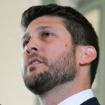 Florida Attorney General James Uthmeier is suing pro-transgender activist groups the American Academy of Pediatrics, the World Professional Association for Transgender Health, and the Endocrine Society. In this picture, Uthmeier with short dark hair and a beard, wearing a suit, looks upward while speaking indoors.