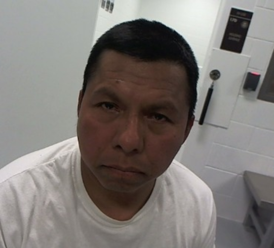 A man with short dark hair wearing a white shirt sits in a room with white walls, gray bench, and a wall-mounted phone visible in the background.