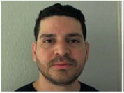A man with short dark hair and a trimmed beard looks directly at the camera, standing against a plain light-colored wall.