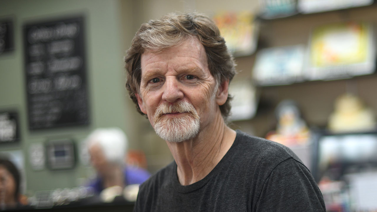 Masterpiece Cakeshop baker Jack Phillips, prosecuted by the Colorado Civil Rights Commission but vindicated by the Supreme Court. A middle-aged man with light brown hair, a beard, and mustache wears a dark t-shirt and sits indoors. Shelves with books and a chalkboard menu are blurred in the background.
