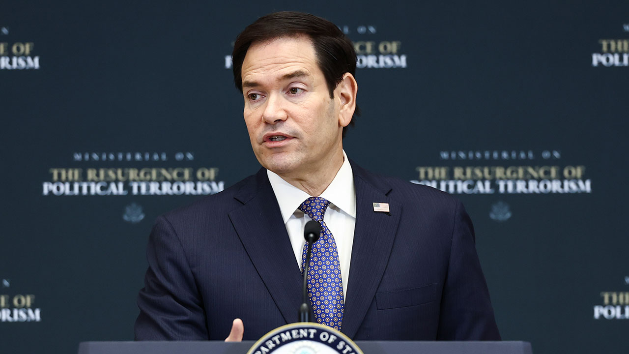 Marco Rubio, wearing a suit and patterned blue tie, speaks at a podium with a sign reading Ministerial on the Resurgence of Political Terrorism and a Department of State seal in front of a dark backdrop. He spoke about leftist terrorism.