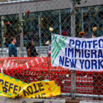 A chain-link fence displays banners reading “PROTECT IMMIGRANT NEW YORKERS” and “DEFEND ASYLEE,” with flowers woven into the fence and people walking behind it.