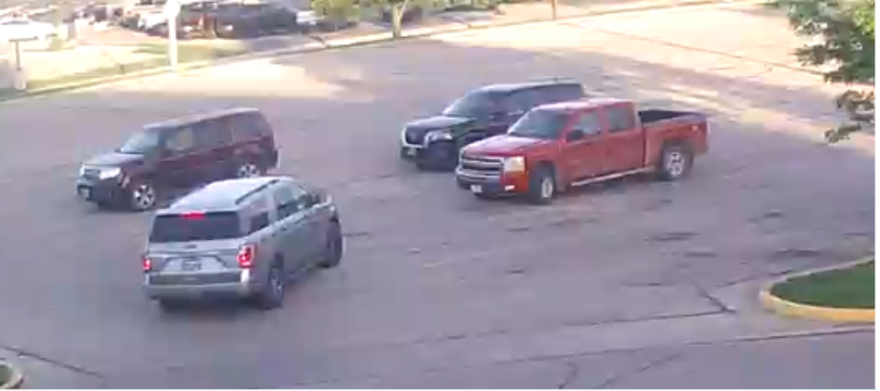 Four vehicles, including a red pickup truck, a silver SUV, a black SUV, and a dark red SUV, are parked or driving in a mostly empty parking lot during daytime.