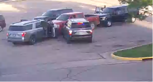 Five vehicles, including a police SUV with flashing lights, are stopped in a parking lot. One vehicle has its drivers door open, and people are standing near two trucks in the background.
