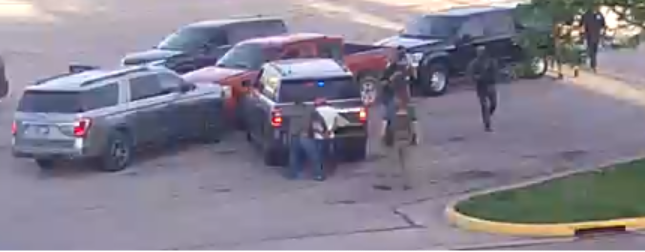 Several vehicles, including SUVs and a pickup truck, are parked close together. Armed individuals in tactical gear stand near the vehicles. The setting appears to be a parking lot near a curb and grassy area.