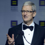 Apple CEO Tim Cook speaks at the Human Rights Campaign. A man in a tuxedo and bow tie speaks at a podium, gesturing with his hands. The background features a logo with yellow equal signs on blue squares and blurred text.