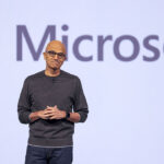 Microsoft CEO Satya Nadella speaks. Microsoft directed Benevity to stop using the Southern Poverty Law Center. Pictured: a person stands on stage in front of a large Microsoft logo projected on a screen, wearing glasses and a dark sweater, with hands clasped.