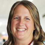 Christian physician assistant Valerie Kloosterman was fired after refusing to use transgender pronouns. A woman with long, wavy brown hair smiles at the camera. She is wearing a white top and is standing indoors with a softly blurred background, possibly a hospital or clinic.