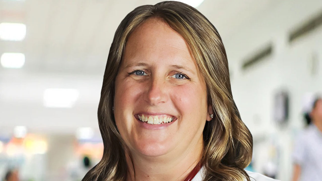 Christian physician assistant Valerie Kloosterman was fired after refusing to use transgender pronouns. A woman with long, wavy brown hair smiles at the camera. She is wearing a white top and is standing indoors with a softly blurred background, possibly a hospital or clinic.