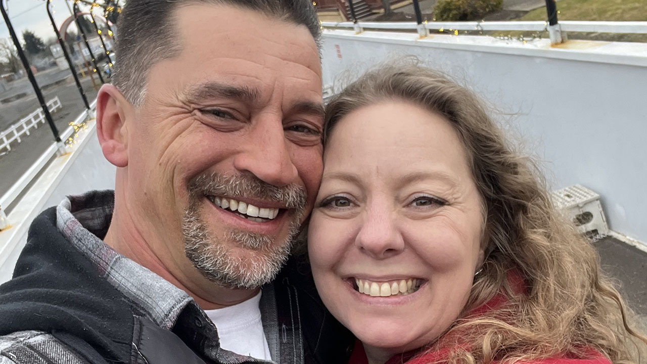 David and Nicole Calaway, parents who have essentially been found guilty of child abuse for causing "mental injury" by not affirming their daughter's transgender identity. A man and woman smiling closely together for a selfie outdoors. The background shows a white fence, pathway, and some lights.