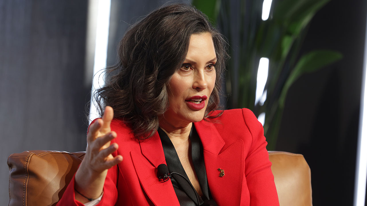 Michigan Gov. Gretchen Whitmer acknowledged that her law against "conversion therapy" violates the First Amendment. Pictured: a woman with wavy dark hair wearing a red blazer sits on a brown chair, gesturing with her hand while speaking. A green plant and blurred background are visible behind her.