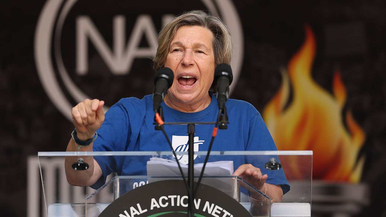 American Federation of Teachers CEO Randi Weingarten. The AFT has endorsed the Democrats' neo-Confederate opposition to President Trump's enforcement of immigration law, a campaign that echoes Nullification. Pictured: a person in a blue shirt speaks passionately at a podium with two microphones. A blurred logo and an image of flames appear in the background.