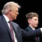 President Donald Trump holds the microphone for Shea Encinas at the Religious Liberty Commission. Pictured: an older man in a suit gestures toward a microphone while a young boy in a suit speaks into it, both standing on a stage with a dark background.