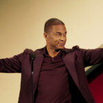 Former CNN host Don Lemon is trying to overturn part of the FACE Act. Pictured: a man in a maroon suit stands at a microphone, smiling and raising both arms. He holds a spiral-shaped award trophy in his left hand against a yellow and red background.