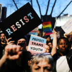 The World Professional Association for Transgender Health just made a key admission. WPATH said its Standards of Care are opinion, not objective fact claims. Pictured: a crowd of protesters holds signs, including RESIST, PROTECT TRANS YOUTH, and Spanish-language messages. People have raised fists, and a trees branches are visible in the background at dusk.