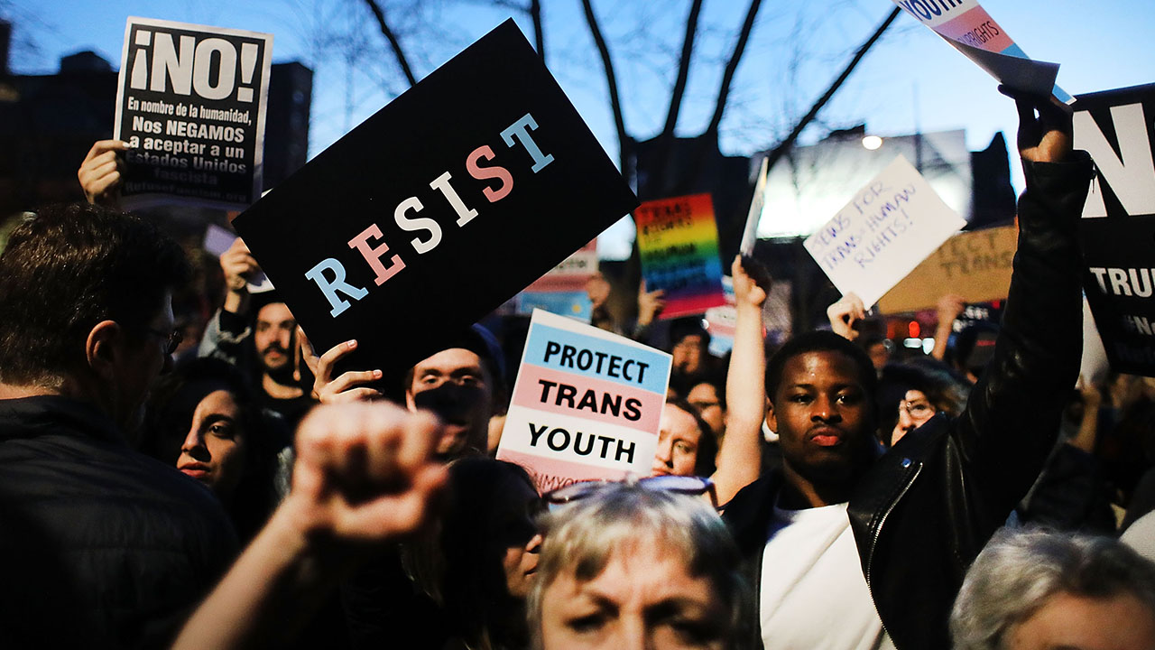 The World Professional Association for Transgender Health just made a key admission. WPATH said its Standards of Care are opinion, not objective fact claims. Pictured: a crowd of protesters holds signs, including RESIST, PROTECT TRANS YOUTH, and Spanish-language messages. People have raised fists, and a trees branches are visible in the background at dusk.