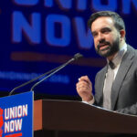 Democratic Socialists of America member and New York City Mayor Zohran Mamdani speaks behind a "union now" poster. Pictured: A man in a suit speaks at a podium with a Union Now sign, in front of a large Union Now banner.