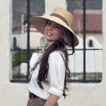 Allison Roberts is suing Portland Public schools over retaliation for her opposition to transgender lessons in school. Pictured: Roberts, a woman in a wide-brimmed straw hat and white blouse smiles while standing outdoors in front of a light-colored wall with iron-barred windows.