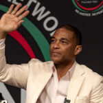Media outlets have come to the defense of former CNN host Don Lemon in the Cities Church case. Pictured: Lemon, a man in a light-colored suit, sits and waves with his right hand at an event, with a black and colorful backdrop behind him.