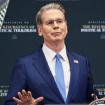 The man who tried to assassinate Treasury Secretary Scott Bessent identifies as transgender and was sentenced to a prison term. Pictured: Bessent, a man in a blue suit and glasses speaks at a podium with his hands raised, standing in front of a backdrop that reads The Resurgence of Political Terrorism.