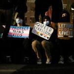 The transgender movement's arguments in court have changed from aggressive promotion to defense. Pictured: Five people sitting, holding protest signs with messages supporting trans and queer rights, such as Protect Trans Kids and Let Trans Kids Live. All are wearing masks, and their faces are partially obscured by shadows.