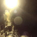 A missile is launched from a naval ship at night, creating a bright burst of light and smoke against the dark sky and ocean. The ships deck and equipment are partially visible under the intense glow.