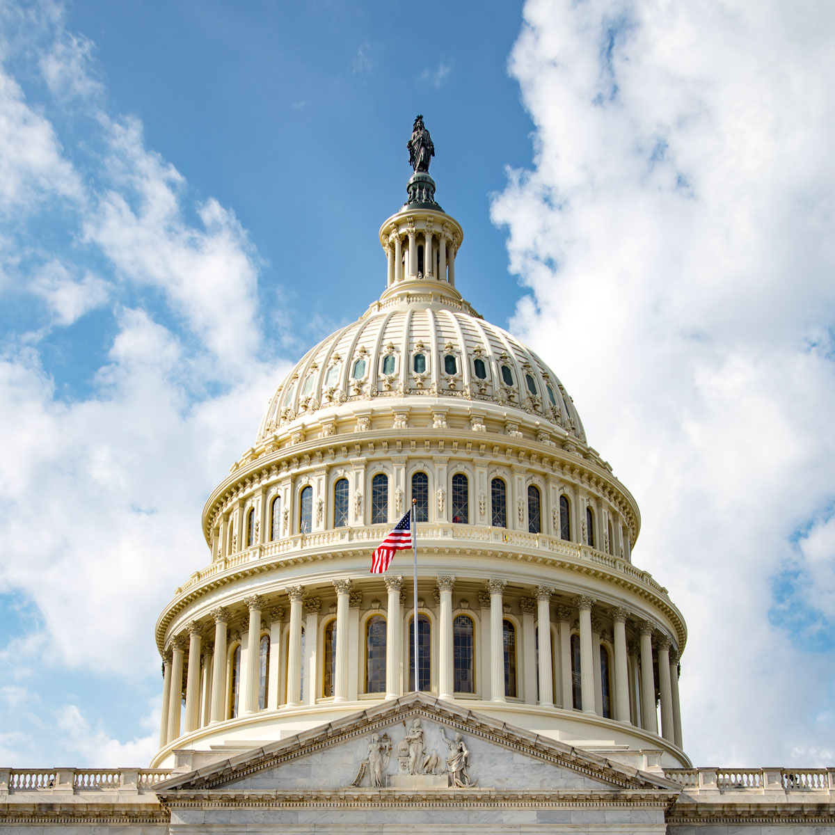 This square crop of the United States Capitol building in Washington, D.C., highlights the detail in the Capitol dome. The backdrop is a blue sky with scattered clouds. An American flag flies in front of the dome.
