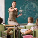 A female teacher sits on a desk in front of a blackboard pointing toward her students.