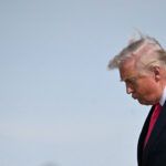 Donald Trump with his head down against a light blue sky.