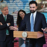New York Mayor Zohran Mamdani stands at a little podium with his arms wide in front of a grafitti-art filled wall. Three associates surround him.