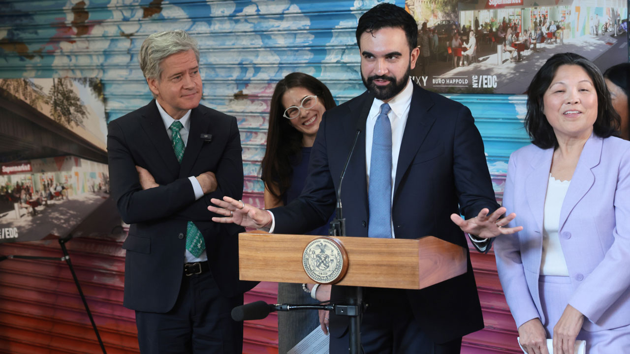 New York Mayor Zohran Mamdani stands at a little podium with his arms wide in front of a grafitti-art filled wall. Three associates surround him.