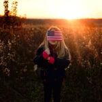 Little girl in a field holds a small American flag in front of her face as the sun sets.