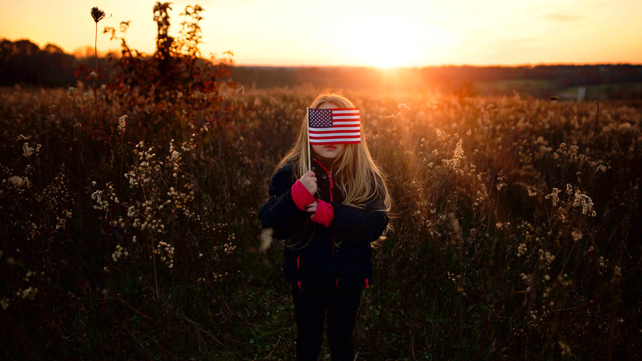 Little girl in a field holds a small American flag in front of her face as the sun sets.
