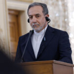 Iranian Foreign Minister Abbas Araghchi stands soberly while wearing headphones at a podium.