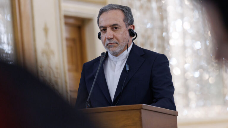 Iranian Foreign Minister Abbas Araghchi stands soberly while wearing headphones at a podium.